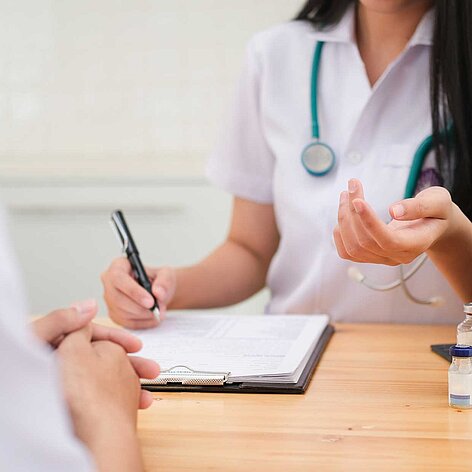 Medizinische Beratung in der Praxis Ärztin im weißen Kittel mit Stethoskop bespricht Informationen mit Patient am Tisch, während sie auf einem Klemmbrett Notizen macht. Ein Impfstoff-Fläschchen steht auf dem Tisch.